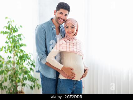 Felici i genitori-a-essere. Felice coppia araba in attesa del bambino, abbracciando e sorridendo alla macchina fotografica, in piedi vicino alla finestra Foto Stock