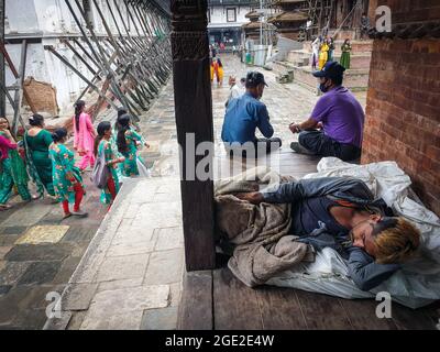 Kathmandu, Bagmati, Nepal. 16 agosto 2021. Un uomo senza dimora dorme sotto un tempio, mentre le donne tornano da un tempio di Shiva dopo aver pregato su Shrawan Somvar in mezzo all'epidemia di COVID-19 a Kathmandu, capitale del Nepal, il 16 agosto 2021. Lunedì o Somvars del mese santo Shrawan sono considerati favorevoli come la gente veloce ed offrire le preghiere al Signore Shiva, specialmente le donne digiunano & pregano per una vita prospera e lunga dei loro mariti o desiderano trovarne una buona. (Credit Image: © Sunil Sharma/ZUMA Press Wire) Credit: ZUMA Press, Inc./Alamy Live News Foto Stock