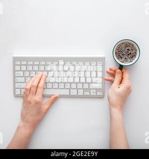 Donna che scrive sulla tastiera del computer e beve caffè sul posto di lavoro. scrivania da lavoro con le mani femminili su sfondo bianco. Foto Stock