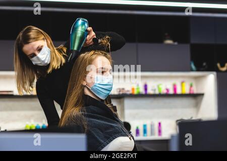 Master donna parrucchiere in una maschera medica asciuga i capelli della ragazza con un asciugacapelli e pettini dopo il lavaggio in un salone di bellezza. Covid-19 pandemia e. Foto Stock