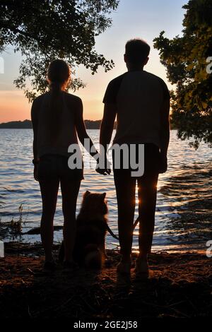 Foto di una coppia che tiene le mani e del cane che si siede tra di loro durante il tramonto in un lago Foto Stock