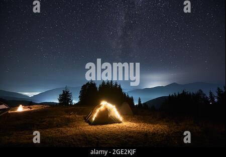 Tenda turistica illuminata da bulbi gialli luminosi su prato verde notte accanto ad un'altra tenda. Campeggio sulla notte stellata. Foresta e sagome di montagna hil Foto Stock