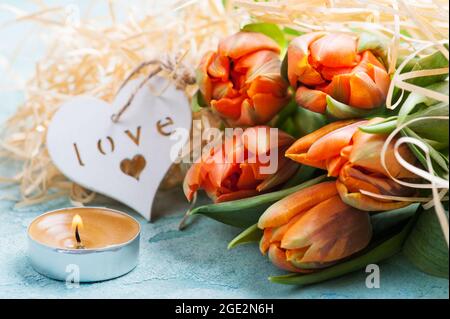 Coppia di tulipani arancio e verde con cuore di legno e candela illuminata su sfondo rustico turchese in cemento. Composizione stile di vita per le vacanze Foto Stock