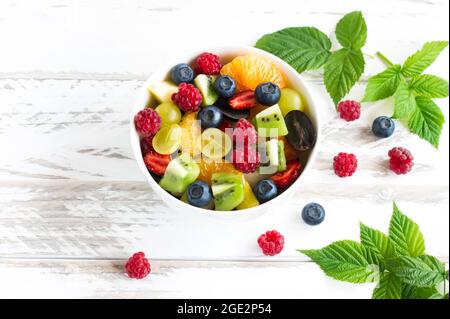 insalata di frutta con banana, kiwi, uva, lamponi, mirtilli in un recipiente su un tavolo di legno bianco Foto Stock