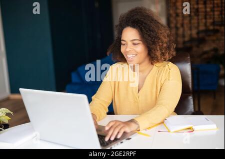 Freelance femminile che lavora con un computer portatile in ufficio. Ottimista afroamericana che scrive sul portatile, conduce corrispondenza con i clienti, invia email, prepara report Foto Stock