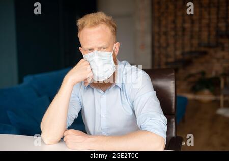 Uomo con testa rossa preoccupato che indossa una maschera medica protettiva a casa, l'uomo pensivo si siede alla scrivania poggiando il mento sulla mano, si sente malinconia. Concetto di autoisolamento Foto Stock
