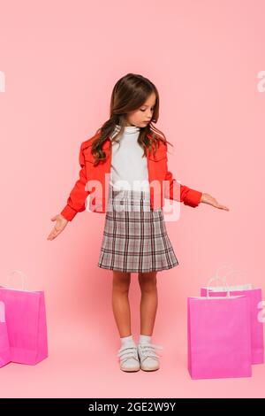 ragazza con giacca arancione e gonna in plaid che punta alle borse della spesa su rosa Foto Stock