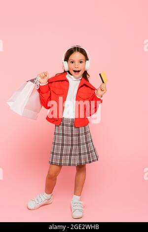 ragazza stupita in cuffie in piedi con carta di credito e borsa per la spesa su rosa Foto Stock