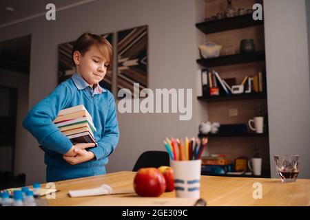 Formazione online a distanza, apprendimento su Internet. Ragazzo di primo grado che studia a casa, tiene un mazzo di libri, preparandosi per la lezione online Foto Stock