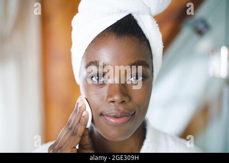 Ritratto di sorridente donna afroamericana in bagno, viso di pulizia con batuffolo di cotone per la cura della pelle Foto Stock