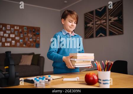Formazione online a distanza, apprendimento su Internet. Ragazzo di primo grado che studia a casa, tiene un mazzo di libri, preparandosi per la lezione online Foto Stock