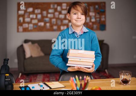 Formazione online a distanza, apprendimento su Internet. Ragazzo di primo grado che studia a casa, tiene un mazzo di libri, preparandosi per la lezione online Foto Stock
