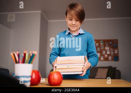 Formazione online a distanza, apprendimento su Internet. Ragazzo di primo grado che studia a casa, tiene un mazzo di libri, preparandosi per la lezione online Foto Stock