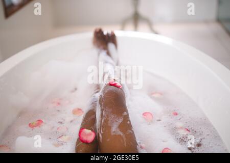 Sezione bassa di donna afroamericana che si rilassa in un bagno di schiuma coccolante con petali di rosa Foto Stock