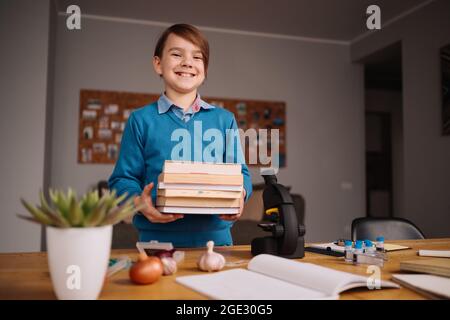 Formazione online a distanza, apprendimento su Internet. Ragazzo di primo grado che studia a casa, tiene un mazzo di libri, preparandosi per la lezione online Foto Stock