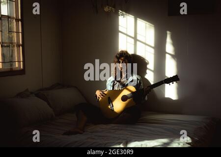 Donna gara mista che suona la chitarra in camera da letto soleggiata Foto Stock