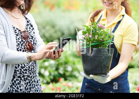 Mani di donna che usano appiccionation moble per controllare l'infroamtion sulla pianta che il responsabile del centro di giardinaggio sta offrendo a lei Foto Stock