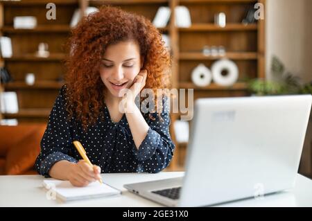 Sorridente giovane donna con capelli rossi ricci parla sullo smartphone utilizzando un computer portatile in ufficio, allegra donna che tiene la telefonata parla con un'espressione positiva del volto, lavora a distanza seduto alla scrivania Foto Stock