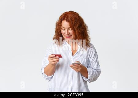 Shopping online, internet banking e concetto di trasferimento di denaro. Ritratto di metà-invecchiata donna rossa guardando la carta di credito, l'acquisto di qualcosa utilizzando Foto Stock