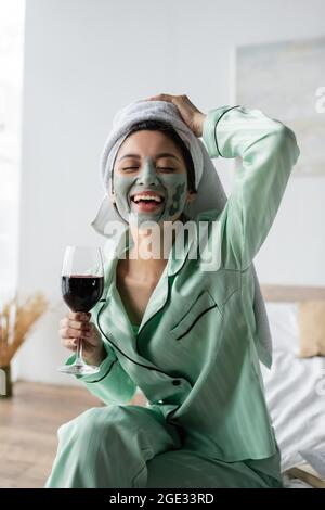 eccitata donna asiatica in maschera di argilla e asciugamano sulla testa tenendo un bicchiere di vino rosso e ridendo con gli occhi chiusi Foto Stock