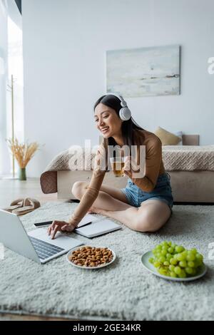 donna asiatica sorridente con una tazza di tè utilizzando un computer portatile sul pavimento in camera da letto Foto Stock