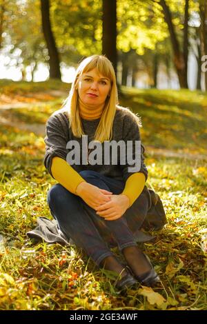 Ritratto di splendida donna di mezza età nel parco autunnale. Bella donna in pullover grigio e stivali in pelle seduta sotto l'albero nel parco autunno con caduto Foto Stock