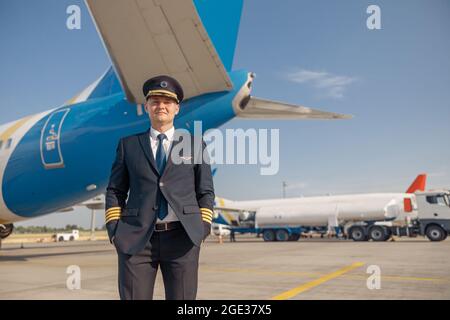 Pilota professionista in uniforme che guarda la macchina fotografica, in piedi di fronte al grande aereo passeggeri pronto per la partenza in aeroporto Foto Stock