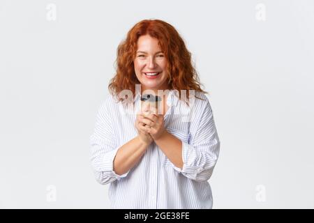 Emozioni, stile di vita e concetto di svago. Bella donna adulta rossa felice in blusa, tenendo una tazza di caffè da asporto e sorridendo gioiosa, godendosi Foto Stock