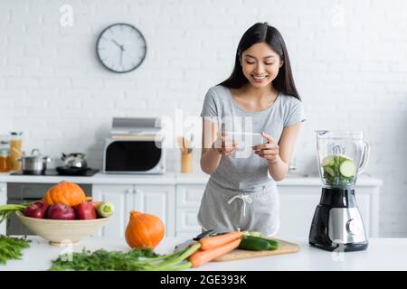 donna asiatica sorridente scattando foto di verdure fresche su smartphone in cucina Foto Stock