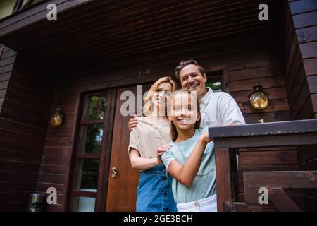 Sorridente ragazza che guarda la macchina fotografica vicino ai genitori abbracciando e casa di vacanza Foto Stock