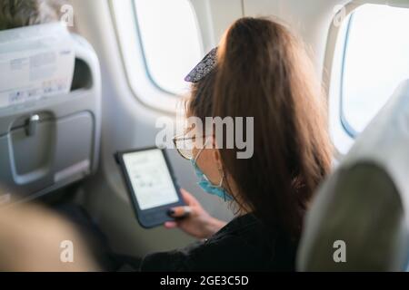 Donna brunetta con maschera sul viso lettura libro elettronico durante il volo su velivolo Foto Stock