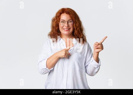 Ritratto di piacevole sorridente donna di mezza età con capelli rossi, indossare occhiali e blusa mostrando pubblicità, cliente di azienda consiglia prodotto o. Foto Stock