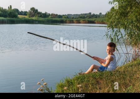 Un bambino piccolo felice seduto sulla riva di uno stagno gioca a pesca con un bastone nelle sue mani. Un giovane pescatore. Bambino, estate e acqua. Divertimento per i bambini in acqua. Tempo di festa. Foto Stock