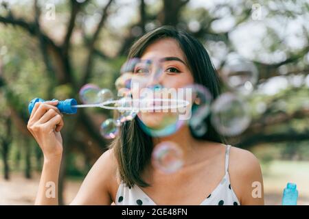 Sud-est asiatico etnico 20s donna soffia bolle d'aria nel parco giovane ragazza felice indossa un abito elegante con gli alberi sullo sfondo. Attività estive all'aperto nel fine settimana - con spazio per fotocopie Foto Stock