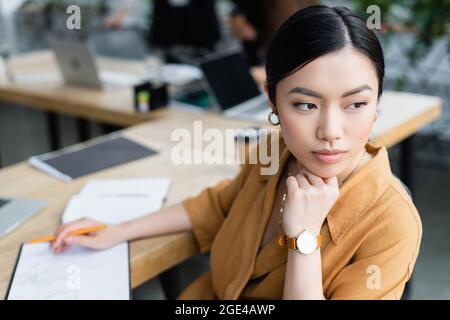 pretty asian designer looking away while thinking near sketch in office Foto Stock