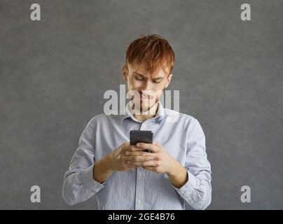 Ritratto di un giovane sorridente che usa un telefono cellulare in piedi sulla parete grigia dello studio Foto Stock