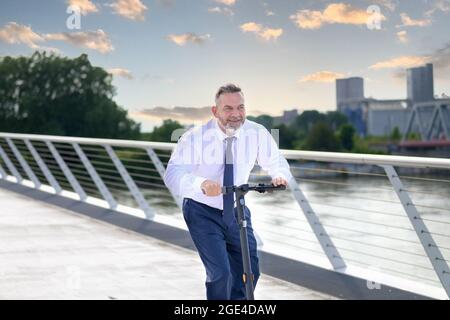 Uomo d'affari che guida uno scooter elettrico su un ponte e un fiume in un ambiente cittadino in una giornata nuvolosa Foto Stock