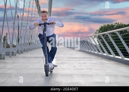 Uomo anziano sportivo felice che guida uno scooter elettrico attraversando un ponte al tramonto con nuvole rosa colorate nel cielo mentre accovacciava sul manubrio Foto Stock