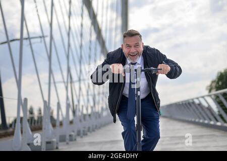 Primo piano di un uomo d'affari senior che guida uno scooter elettrico attraverso un ponte al tramonto Foto Stock