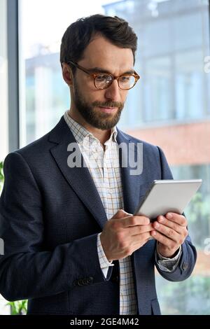 Uomo d'affari serio e maturo che utilizza un dispositivo tablet in piedi in ufficio. Foto Stock