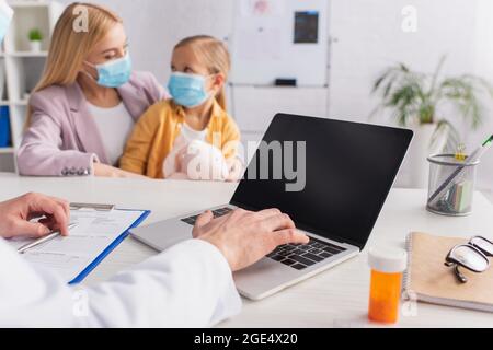Medico usando il laptop vicino pillole e madre con la figlia in maschere mediche su sfondo offuscato Foto Stock