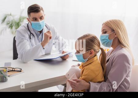 Madre in maschera medica guardando la ragazza con il giocattolo morbido vicino pediatra Foto Stock