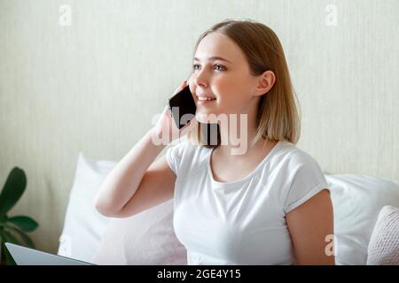 Giovane donna sorridente che parla via telefono in camera da letto. Conversazione con smartphone al mattino a casa. Felice giovane bionda caucasica comunica Foto Stock