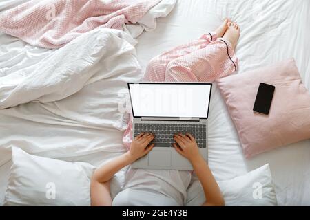 La giovane donna in pigiama si trova a letto utilizzando un computer portatile con un modello di mockup vuoto sullo schermo per il design e lo smartphone. Lavoro remoto da casa Foto Stock
