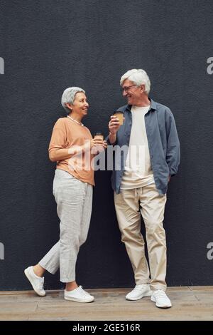 Ritratto verticale a lunghezza intera di una coppia matura moderna che si guarda l'un l'altro e sorridente mentre si trova in piedi da una parete grigia Foto Stock
