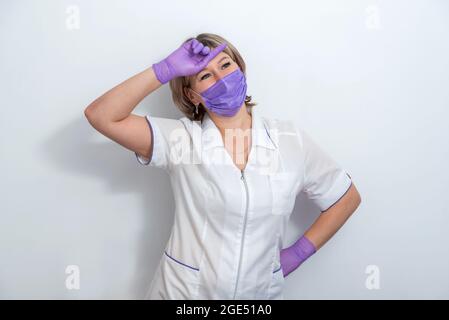 Infermiera in una uniforme medica in piedi con la mano sollevata alla fronte e indossando camice bianco, maschera protettiva e guanti. Isolato su bianco Foto Stock