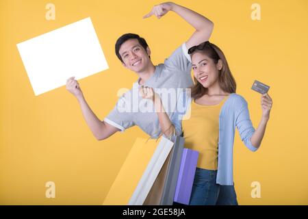 Coppia asiatica che tiene il cartellone vuoto con carta di credito e borse per la spesa isolati su sfondo giallo Foto Stock