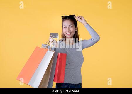 Donna asiatica felice con shopping borse e carta di credito sorridente su sfondo giallo, spazio vuoto Foto Stock