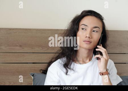 Ritratto di un giovane serio seduto a letto e che parla al telefono con gli amici Foto Stock