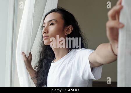 Bel giovane uomo in maglietta bianca che apre le tende nella sua camera da letto e guarda all'esterno Foto Stock